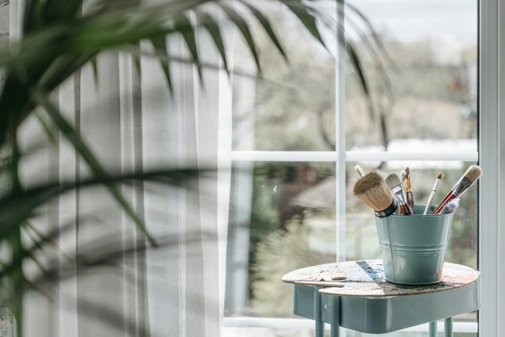 A bucket of art brushes on a table by a sunny window, inspiring creativity.